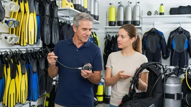 A man and a woman inside a well-lit scuba diving equipment shop, examining a regulator and a buoyancy compensator with other 